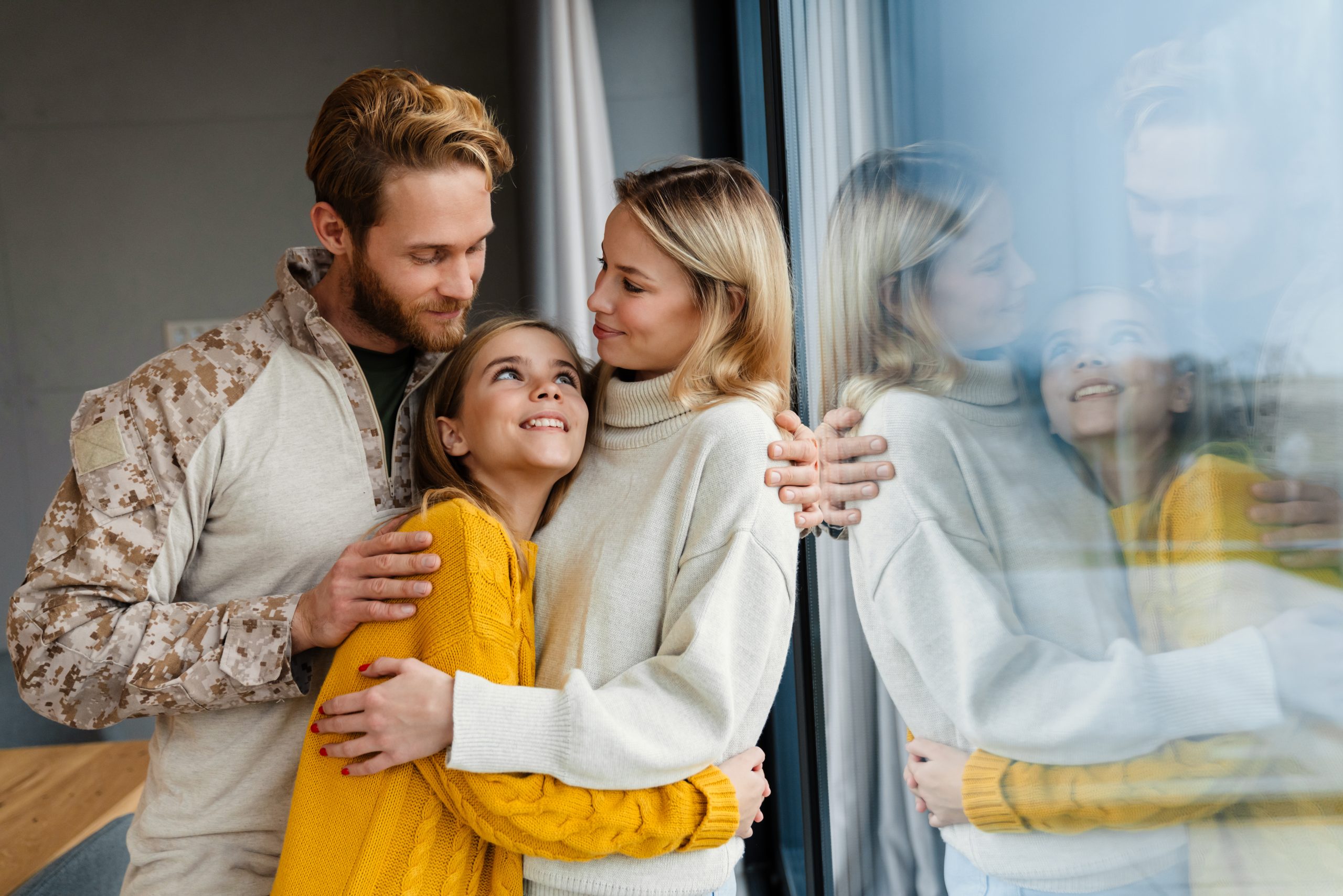 Dad in a camouflage jacket hugging his wife and young daughter by a large glass window, all smiling at each other.